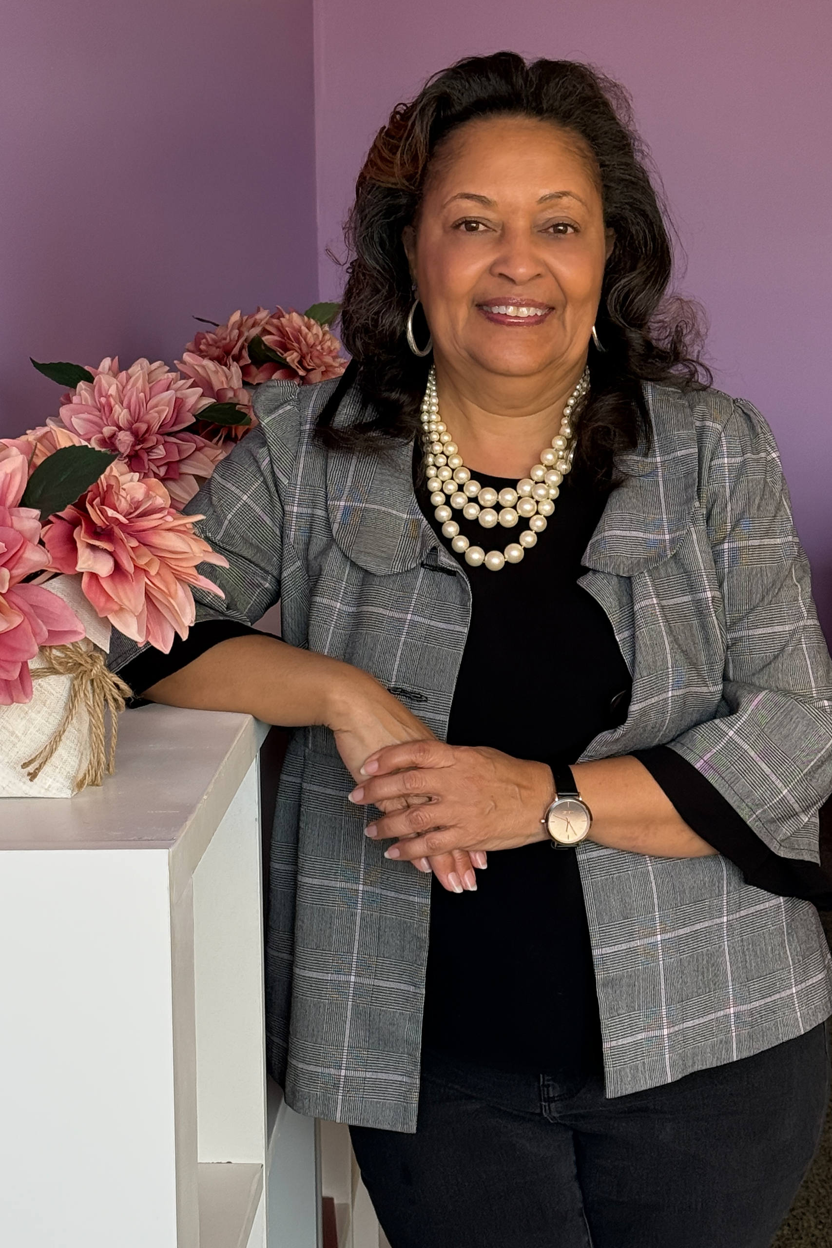 Professional woman smiling while leaning on a white shelf, wearing a gray plaid blazer over a black top with a layered pearl necklace and watch, standing in front of a soft purple wall with pink flowers beside her.